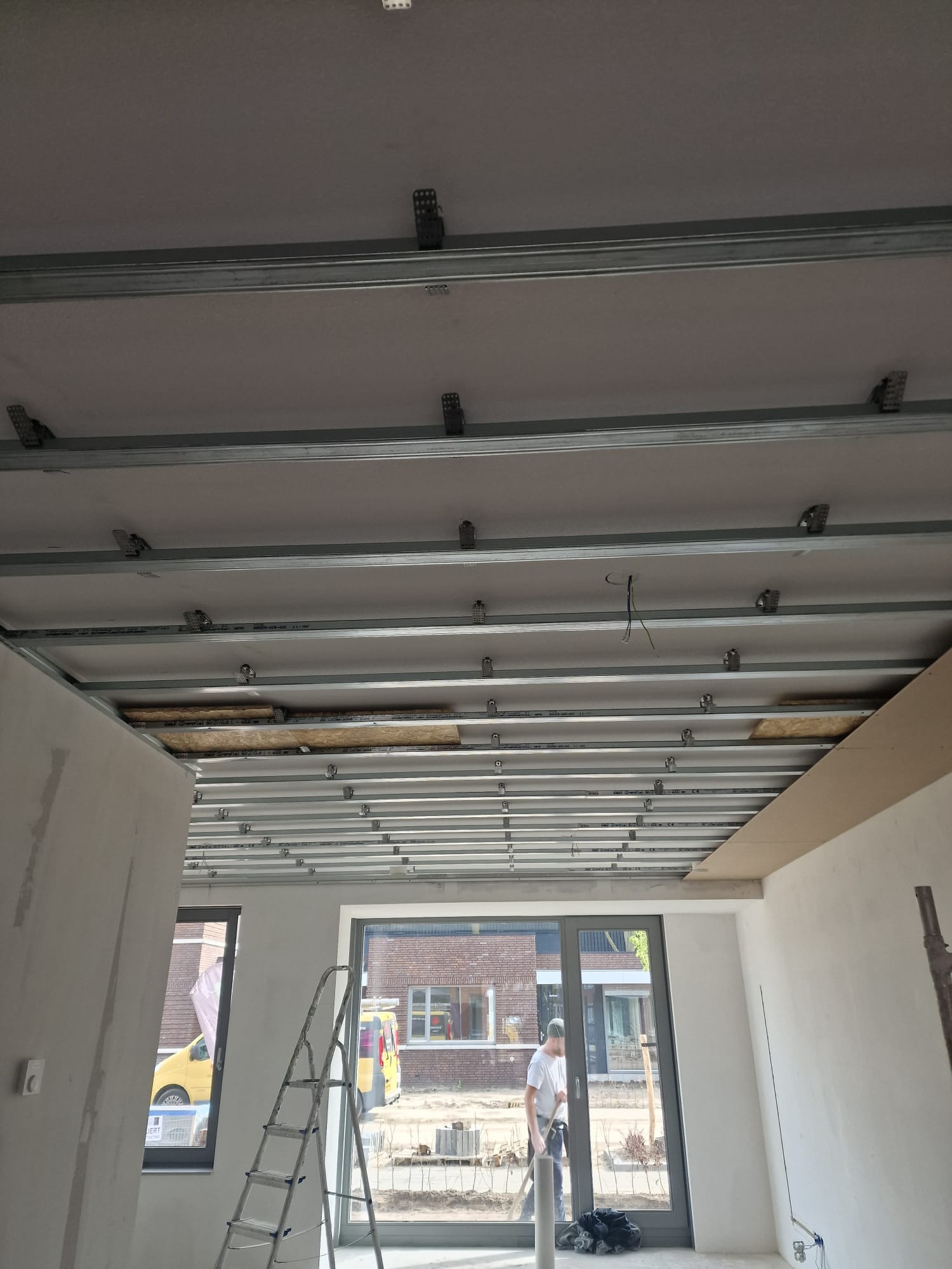 Montaż metalowej konstrukcji sufitu podwieszanego z widocznymi elementami mocującymi, częściowo zakrytej płytami, w tle okno z widokiem na ulicę i pracownik.