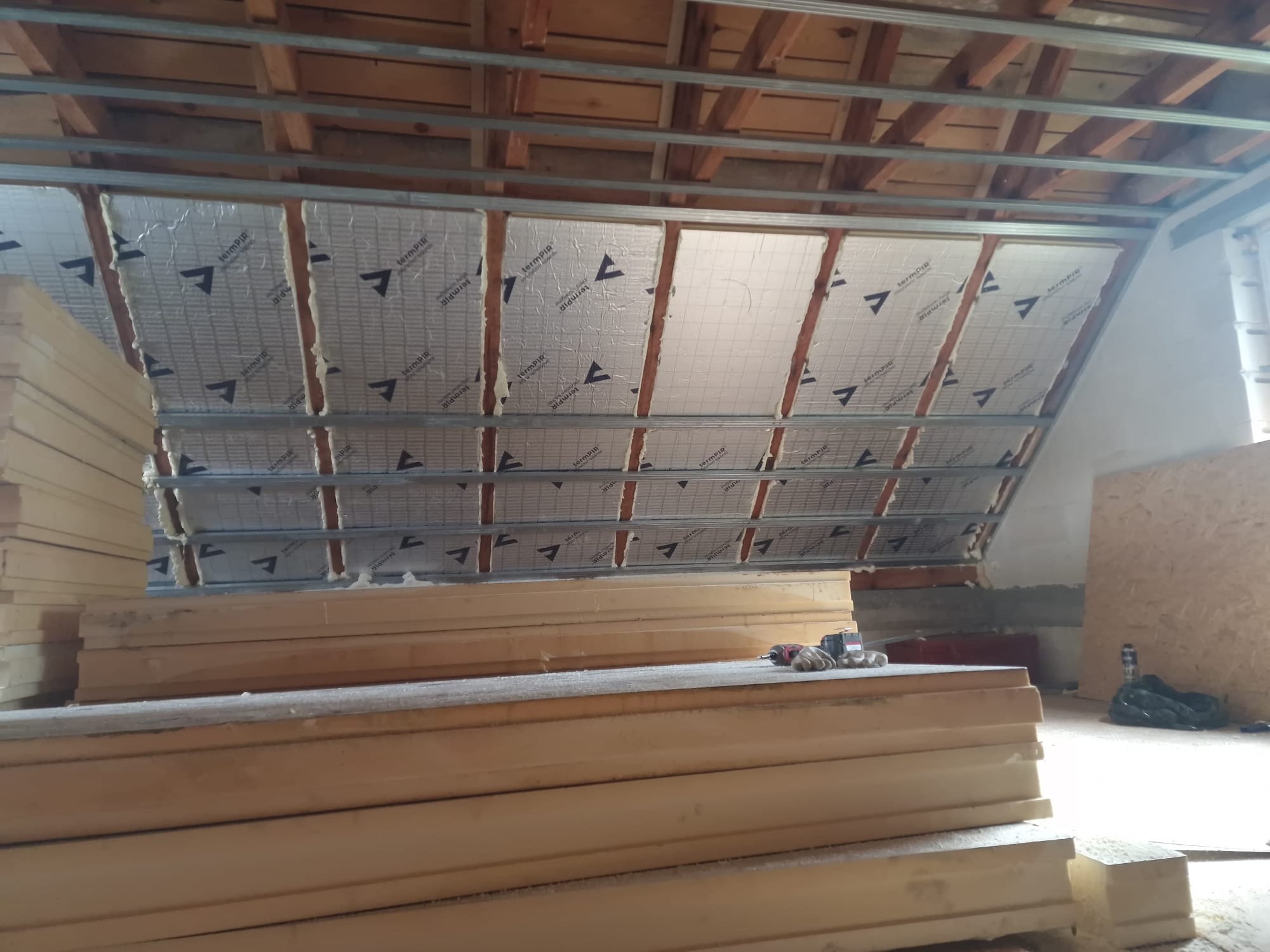 Poddasze w trakcie izolacji termicznej, widoczne metalowe profile, wełna mineralna z folią aluminiową i stos płyt izolacyjnych z pianki poliuretanowej.