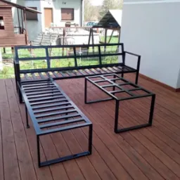 Metalowy komplet mebli ogrodowych: ławka z oparciem, długa ławka bez oparcia i stolik kawowy, wykonane z czarnych profili, na drewnianym tarasie.