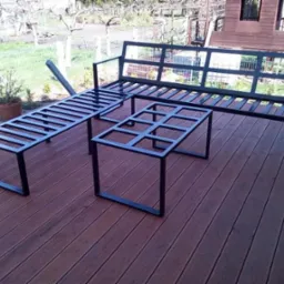 Czarny, metalowy komplet mebli ogrodowych: sofa, ławka i stolik kawowy, ustawione na drewnianym tarasie z widokiem na ogród.