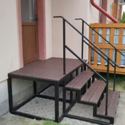 Zewnętrzne schody z podestem wykonane z kompozytu drewnopodobnego na metalowej konstrukcji, z czarną, prostą balustradą, prowadzące do drzwi wejściowych.