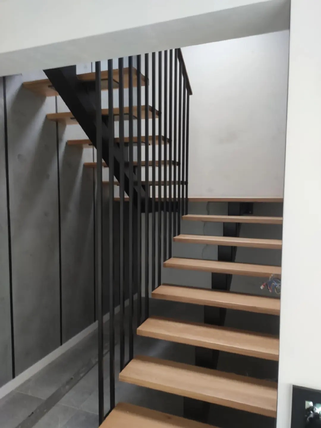 Nowoczesne schody z drewnianymi stopniami i czarną metalową balustradą z pionowych prętów w minimalistycznym wnętrzu.