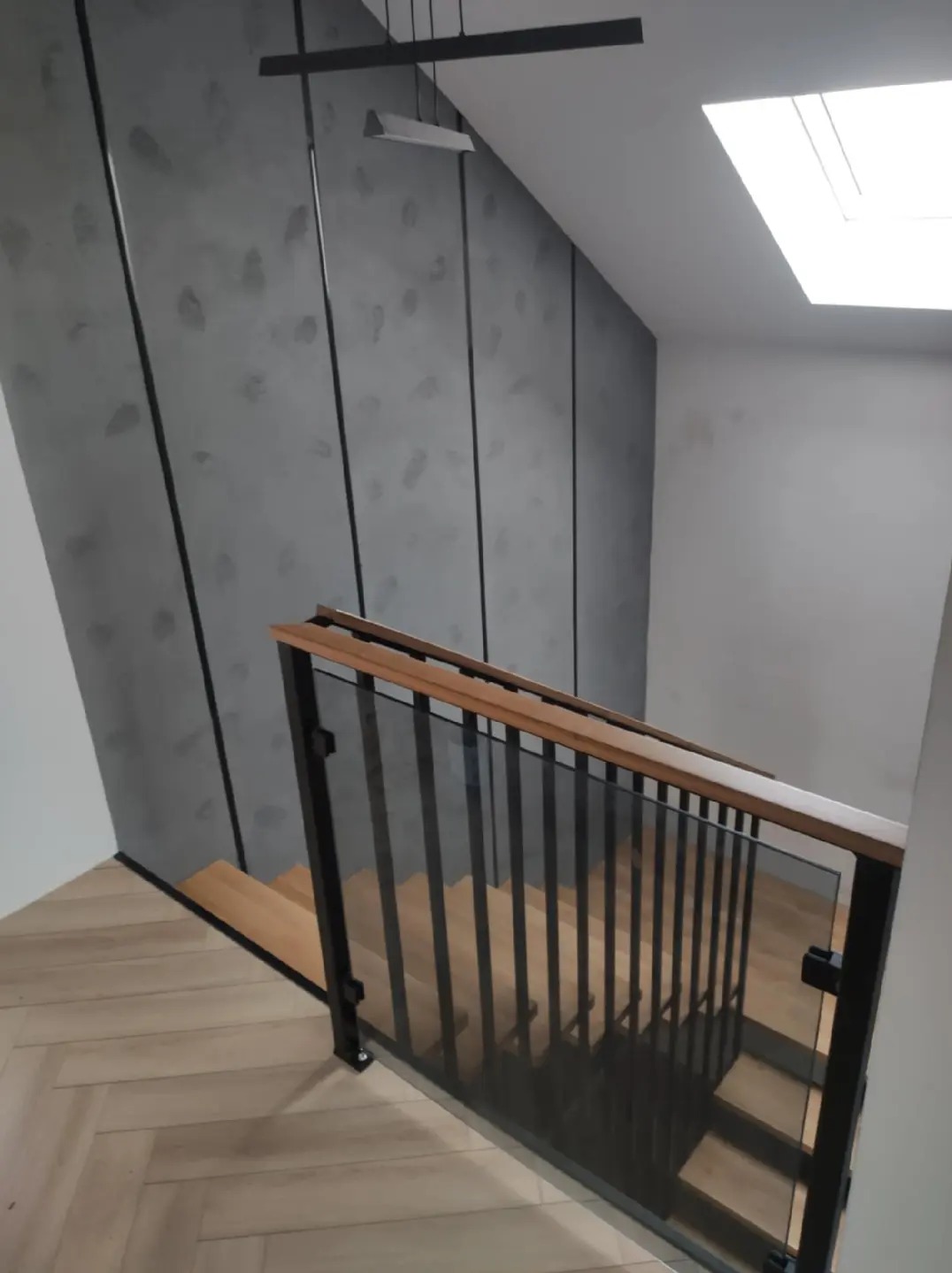 Nowoczesne schody drewniane z czarnymi metalowymi balustradami i szklanymi panelami, widok z góry na dół, parkiet ułożony w jodełkę, szara ściana z dekoracyjnymi czarnymi listwami, oświetlenie...