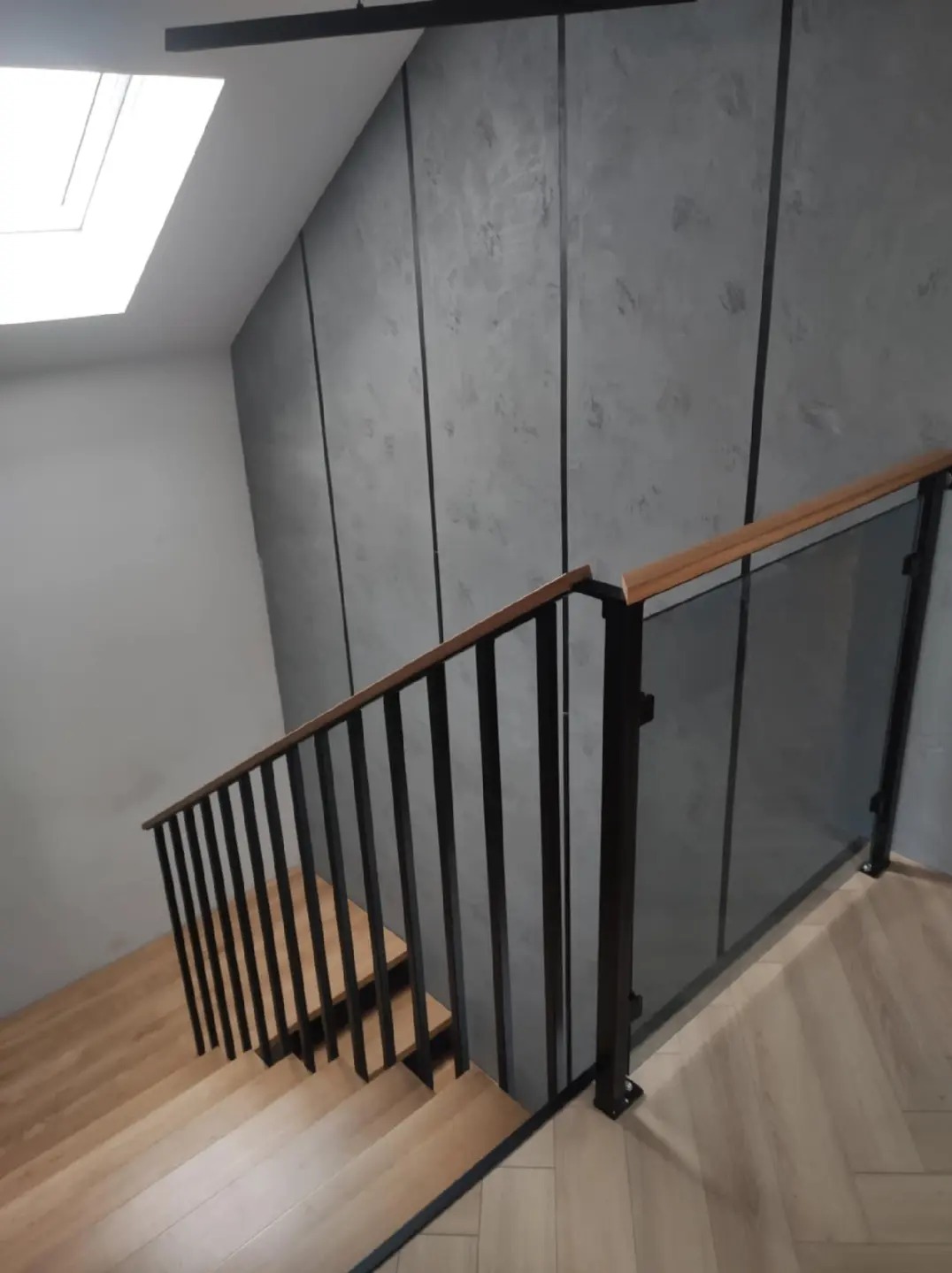 Drewniane schody z czarną metalową balustradą i szklanym panelem, widok z góry, szara ściana z pionowymi panelami, oświetlenie górne.