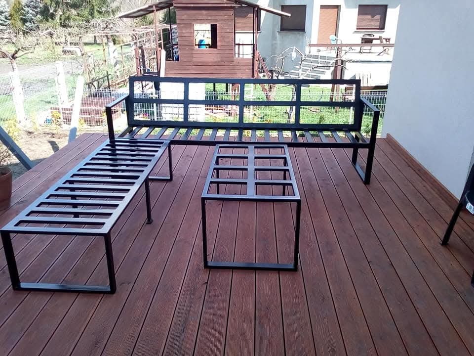 Metalowy zestaw mebli ogrodowych: ławka z oparciem, stolik kawowy i długa ławka bez oparcia, na drewnianym tarasie przy domu.