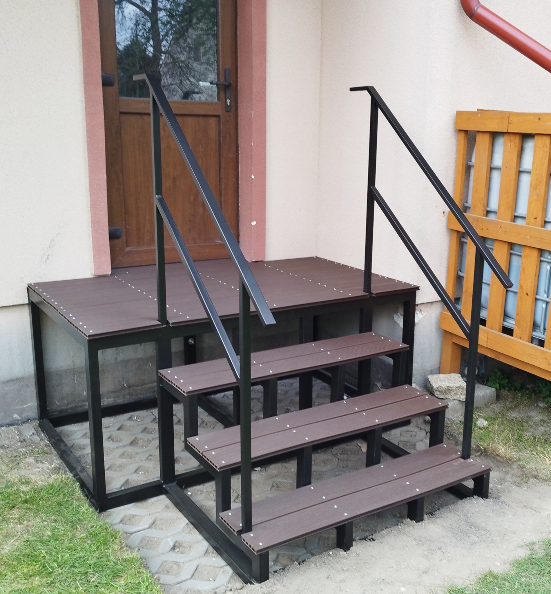 Zewnętrzne schody z podestem, konstrukcja metalowa w kolorze czarnym, wykończona deskami kompozytowymi w odcieniach brązu, widoczne mocowania na śruby.