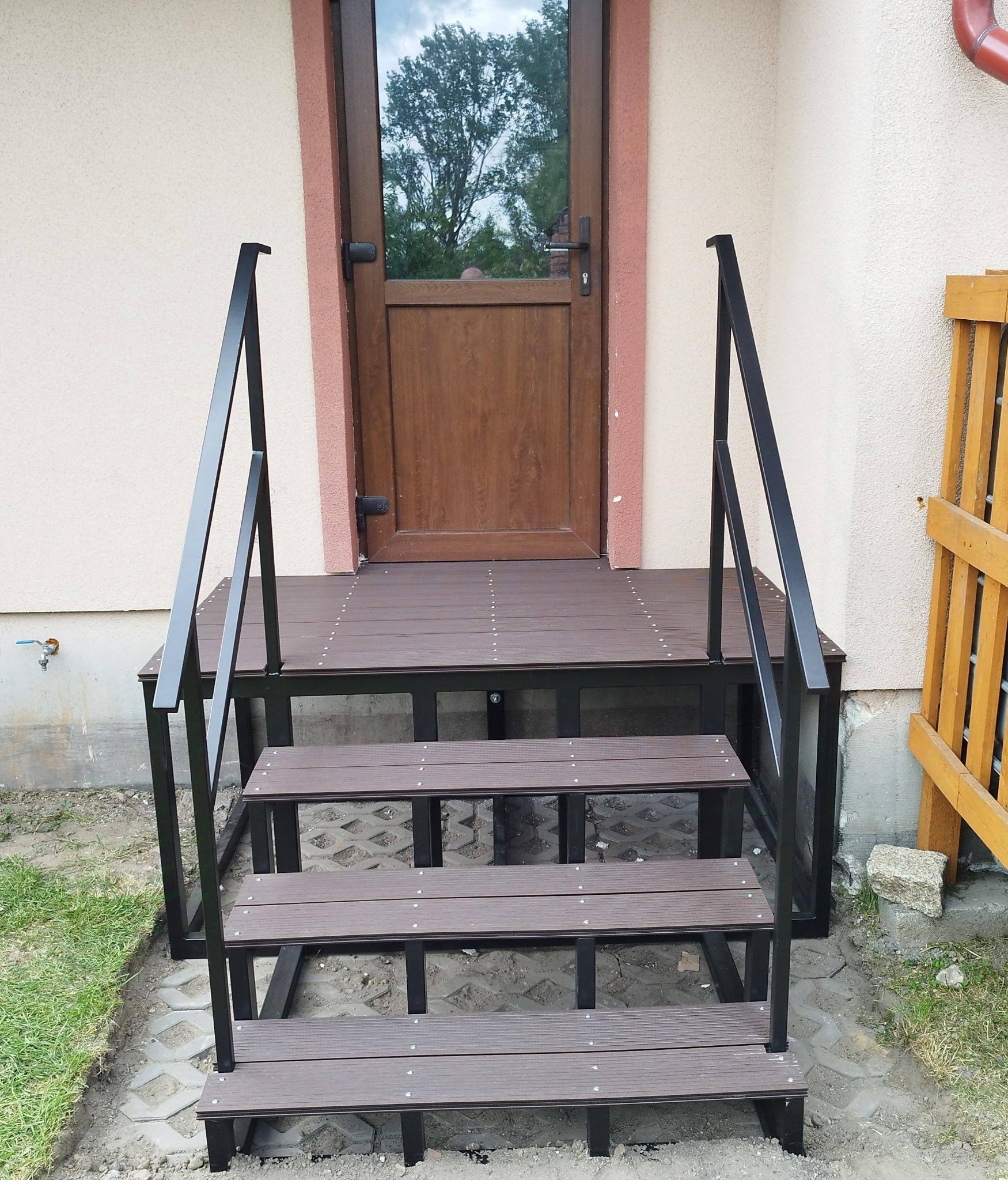 Zewnętrzne schody z kompozytu drewnopodobnego z czarną metalową konstrukcją i poręczami, prowadzące do drzwi wejściowych.