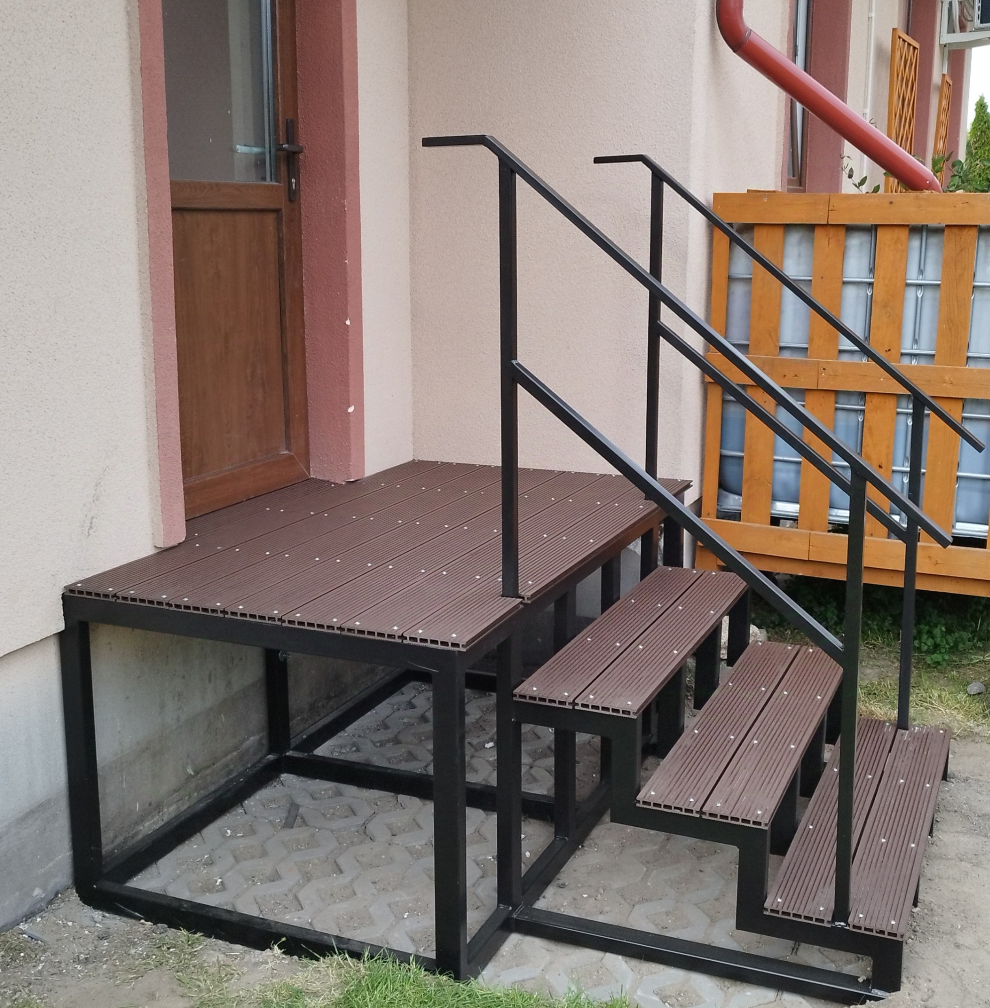 Zewnętrzne schody z podestem wykonane z kompozytu drewnopodobnego na metalowej konstrukcji, z czarną, prostą balustradą, prowadzące do drzwi wejściowych.