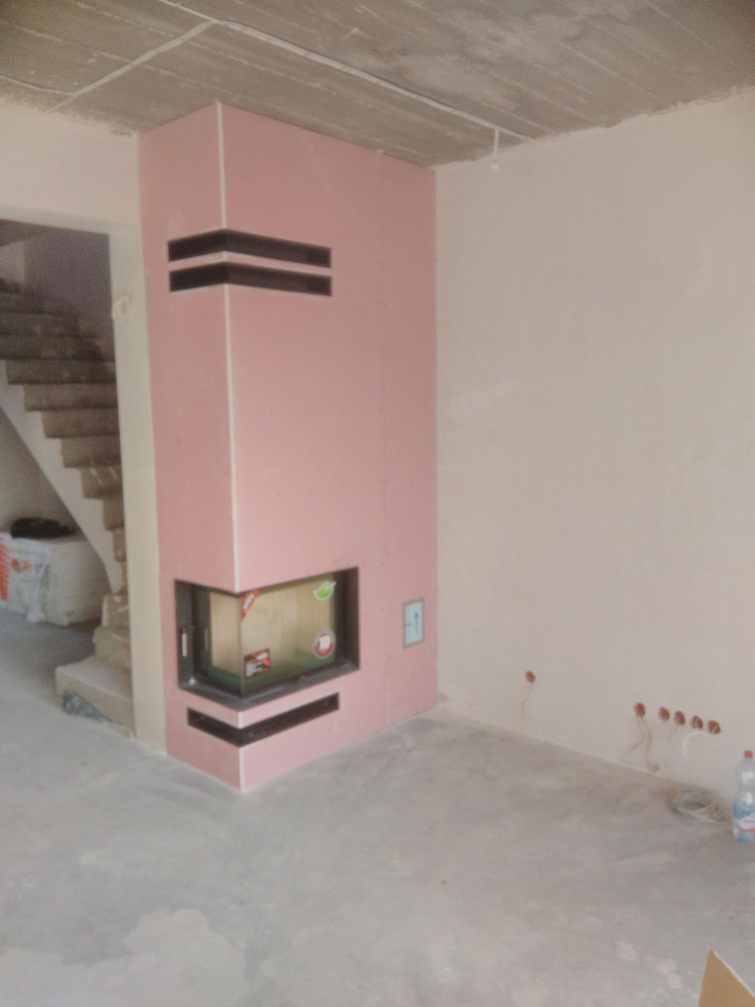 Wnętrze w Bytomiu w trakcie wykańczania z obudowanym wkładem kominkowym w kolorze różowym z czarnymi akcentami, widoczne schody i surowy betonowy strop.