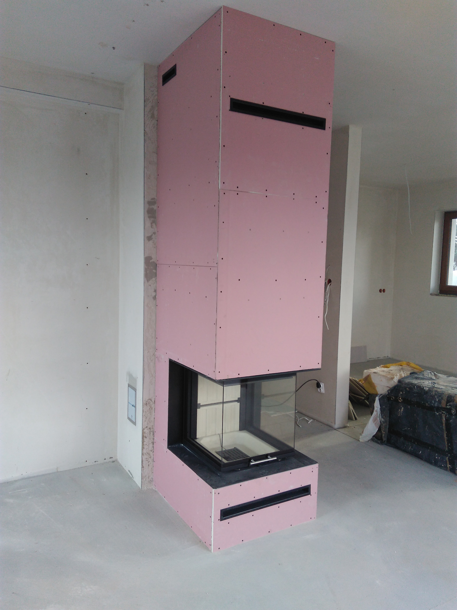 Narożny kominek obudowany płytami gipsowo-kartonowymi w surowym stanie, z czarnym wkładem i szybą, w jasnym pomieszczeniu w trakcie wykończenia.