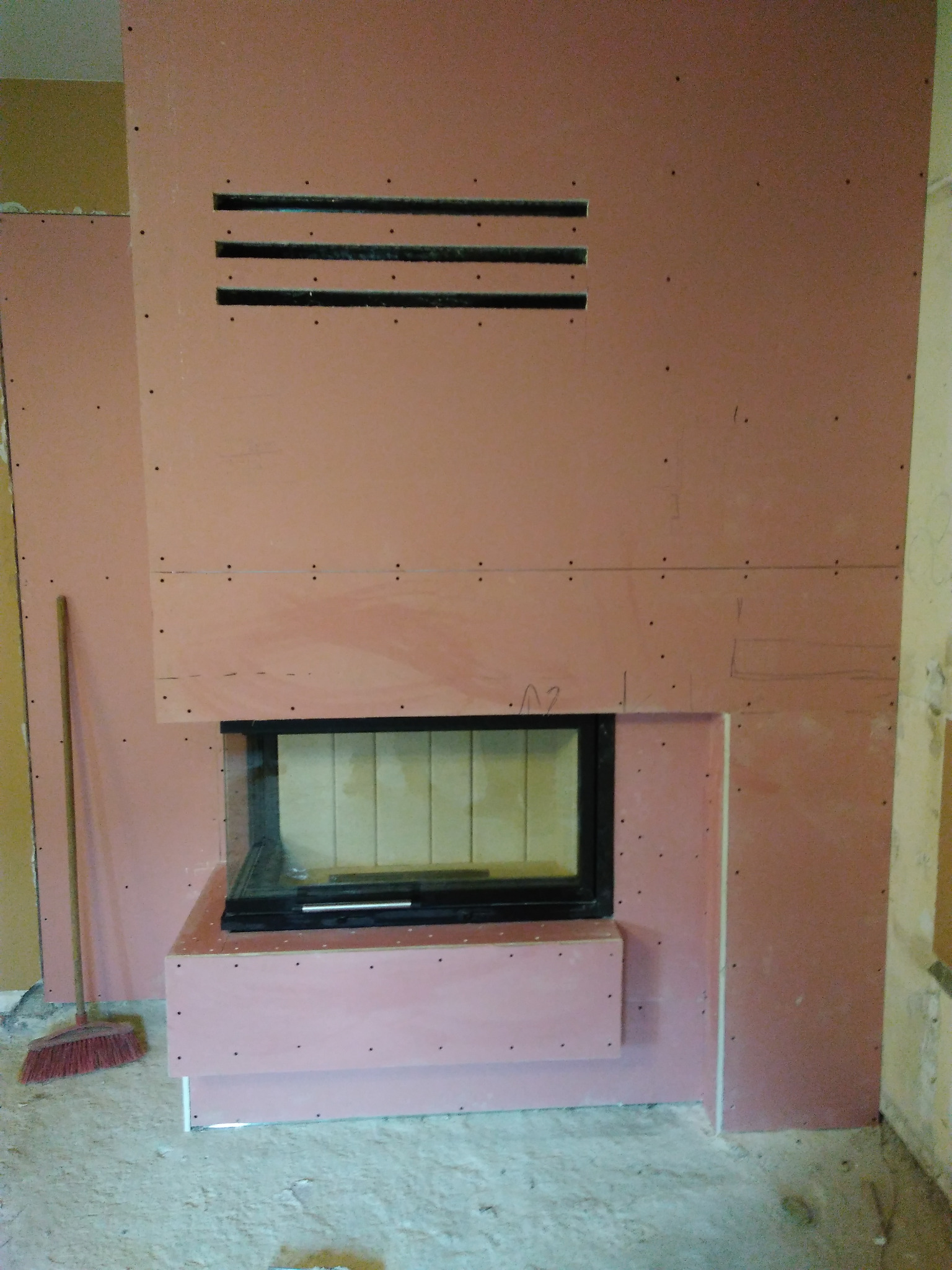 Montaż kominka w obudowie z płyt gipsowych, widoczne otwory wentylacyjne. Etap prac wykończeniowych, z boku stoi miotła.