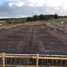PÓŁNOC CHYCZEWSKA SPÓŁKA KOMANDYTOWA - Siatka zbrojeniowa ułożona w przygotowanym wykopie pod fundamenty, widoczne szalunki i teren budowy w tle.
