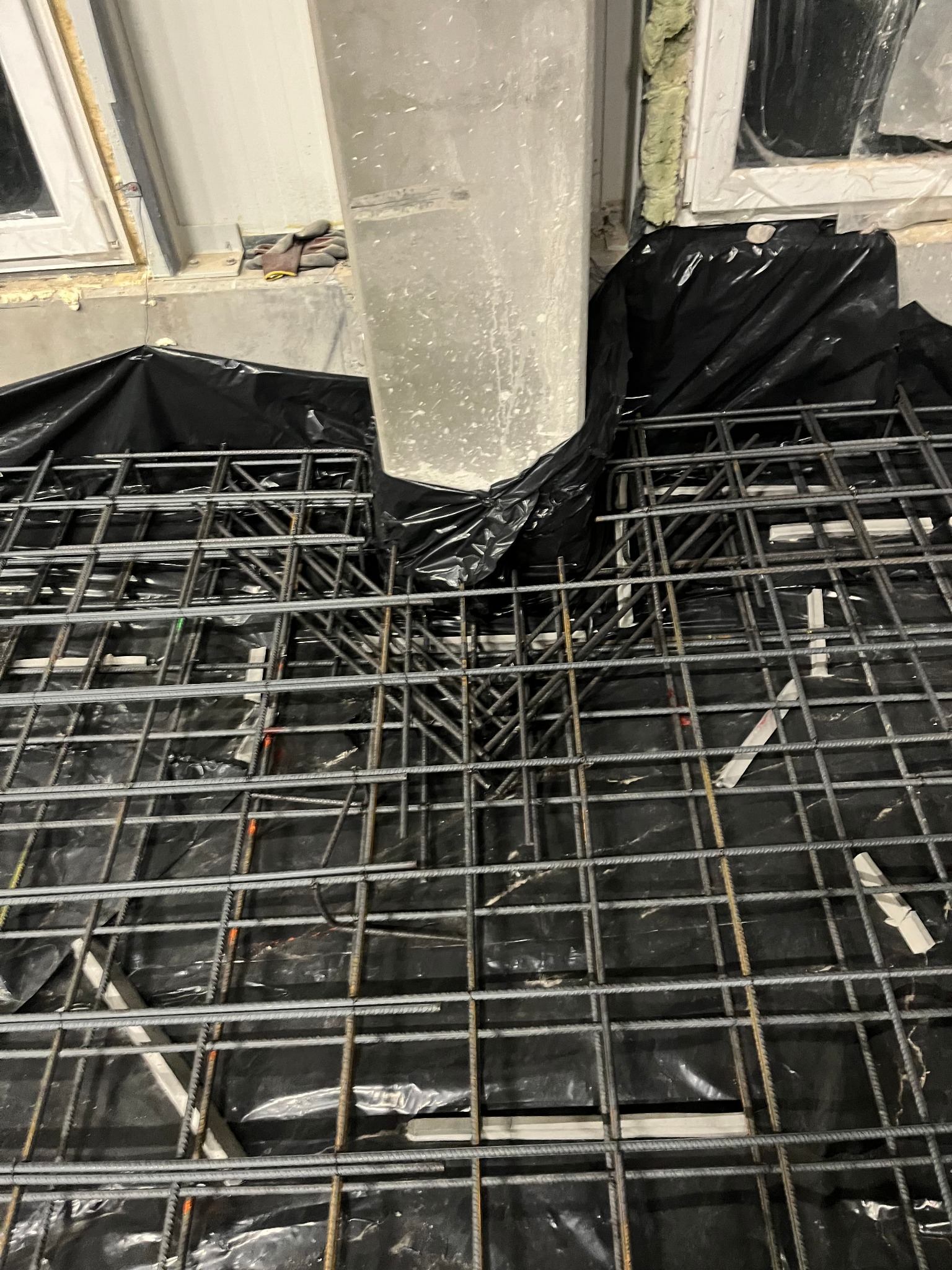 Zbrojenie fundamentu z siatki stalowej, zabezpieczone folią przed wylaniem betonu, widoczny słup konstrukcyjny i robocze rękawice na parapecie okna.