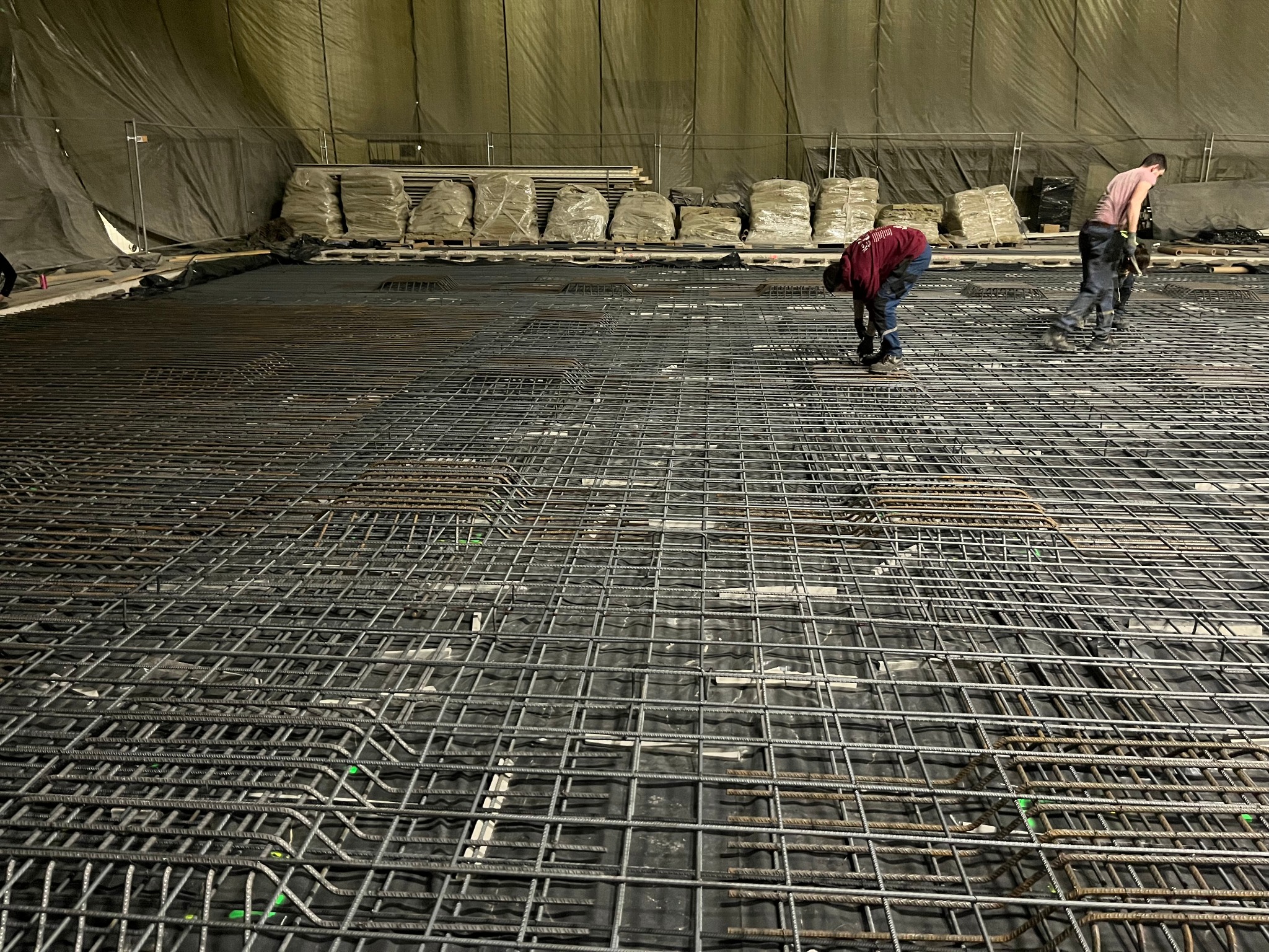 Dwóch zbrojarzy pracuje nad siatką zbrojeniową na czarnej folii, w tle worki z materiałami budowlanymi i zasłonięta ściana.