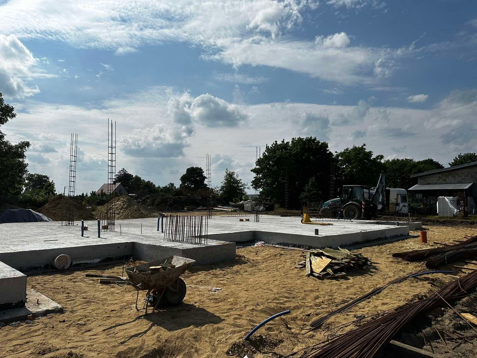 Płyta fundamentowa z wystającymi prętami zbrojeniowymi, taczka i koparka w tle na piaszczystym terenie budowy w słoneczny dzień.