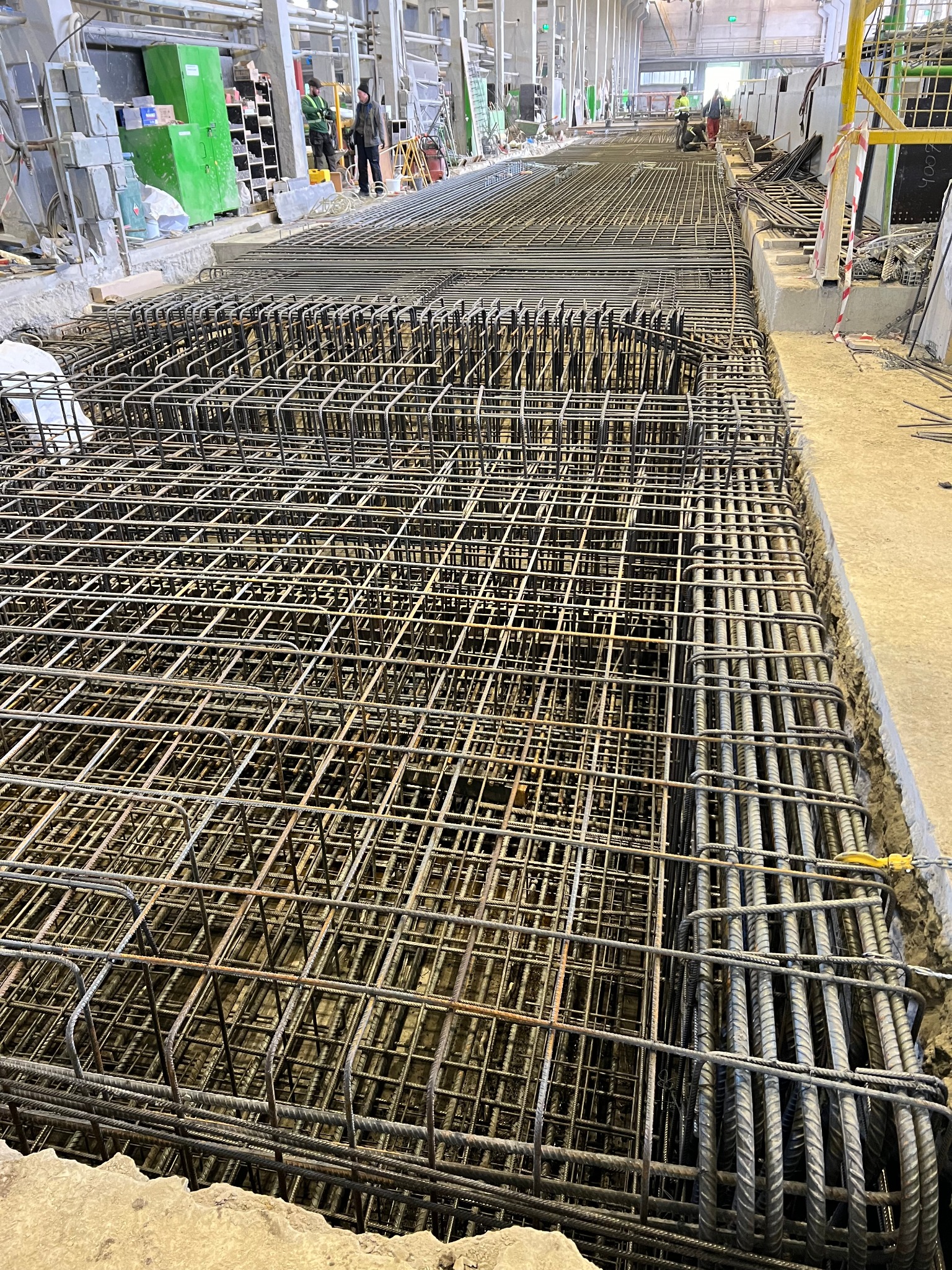 Siatka zbrojeniowa fundamentów, widok z góry, zarys wykopu, na drugim planie pracownicy budowlani i sprzęt.