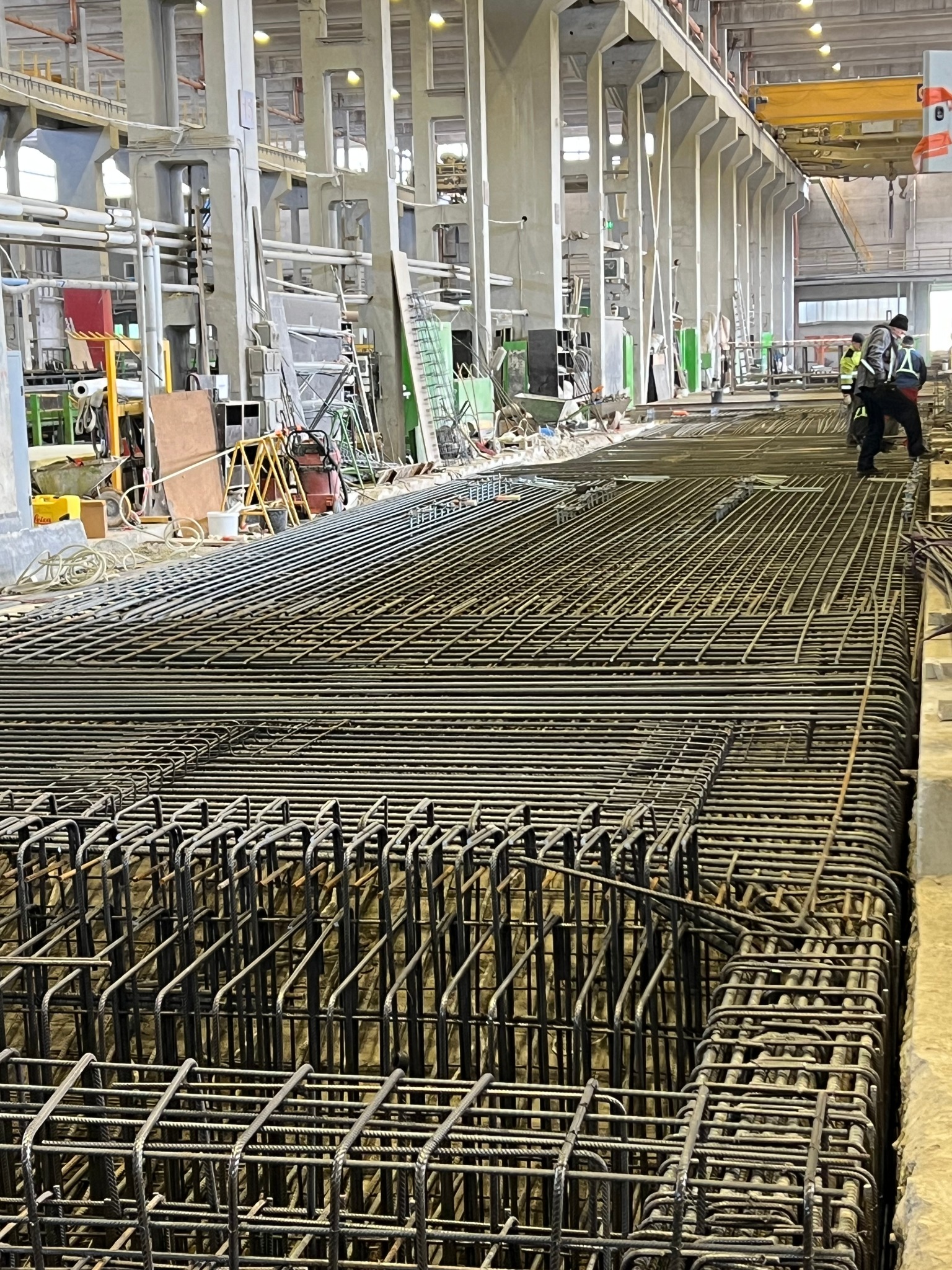 Siatka zbrojeniowa ułożona w dużej hali przemysłowej, przygotowana do zalania betonem pod fundamenty, z widocznymi pracownikami w tle.