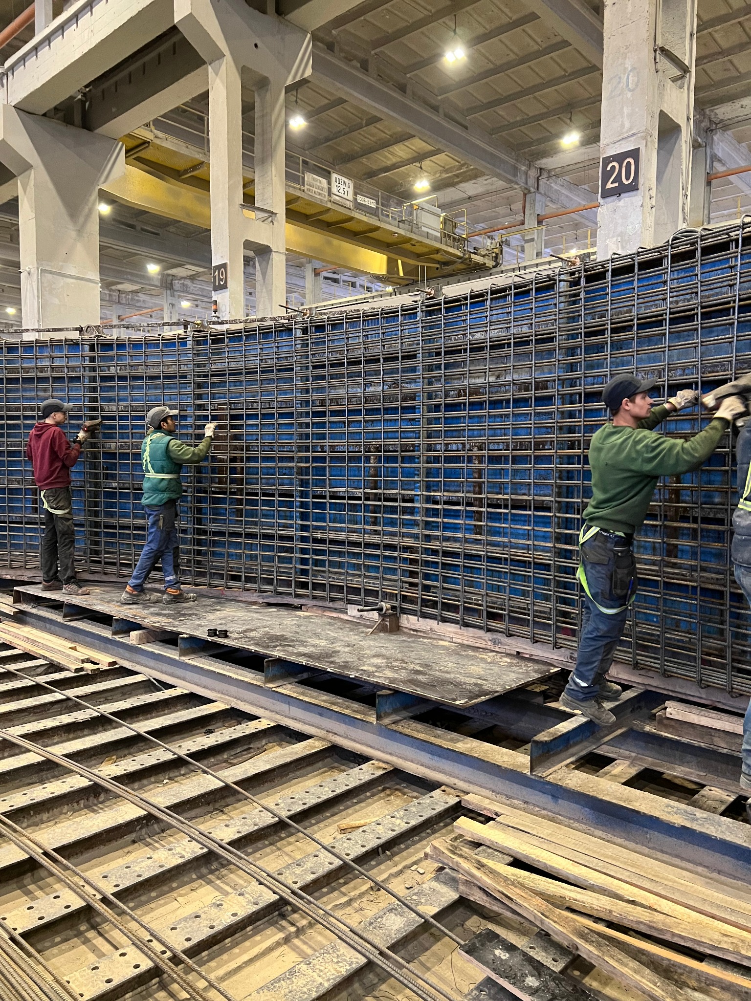 Pracownicy budowlani montują zbrojenie w szalunku na tle hali produkcyjnej z suwnicą i numerowanymi słupami.