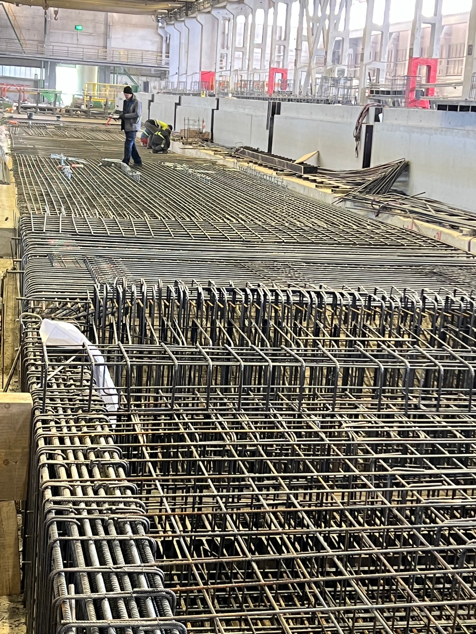 Siatka zbrojeniowa ułożona na budowie, dwóch pracowników w tle, betonowe ściany w oddali.
