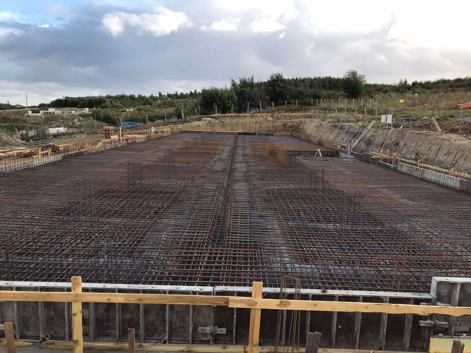 Siatka zbrojeniowa ułożona w przygotowanym wykopie pod fundamenty, widoczne szalunki i teren budowy w tle.