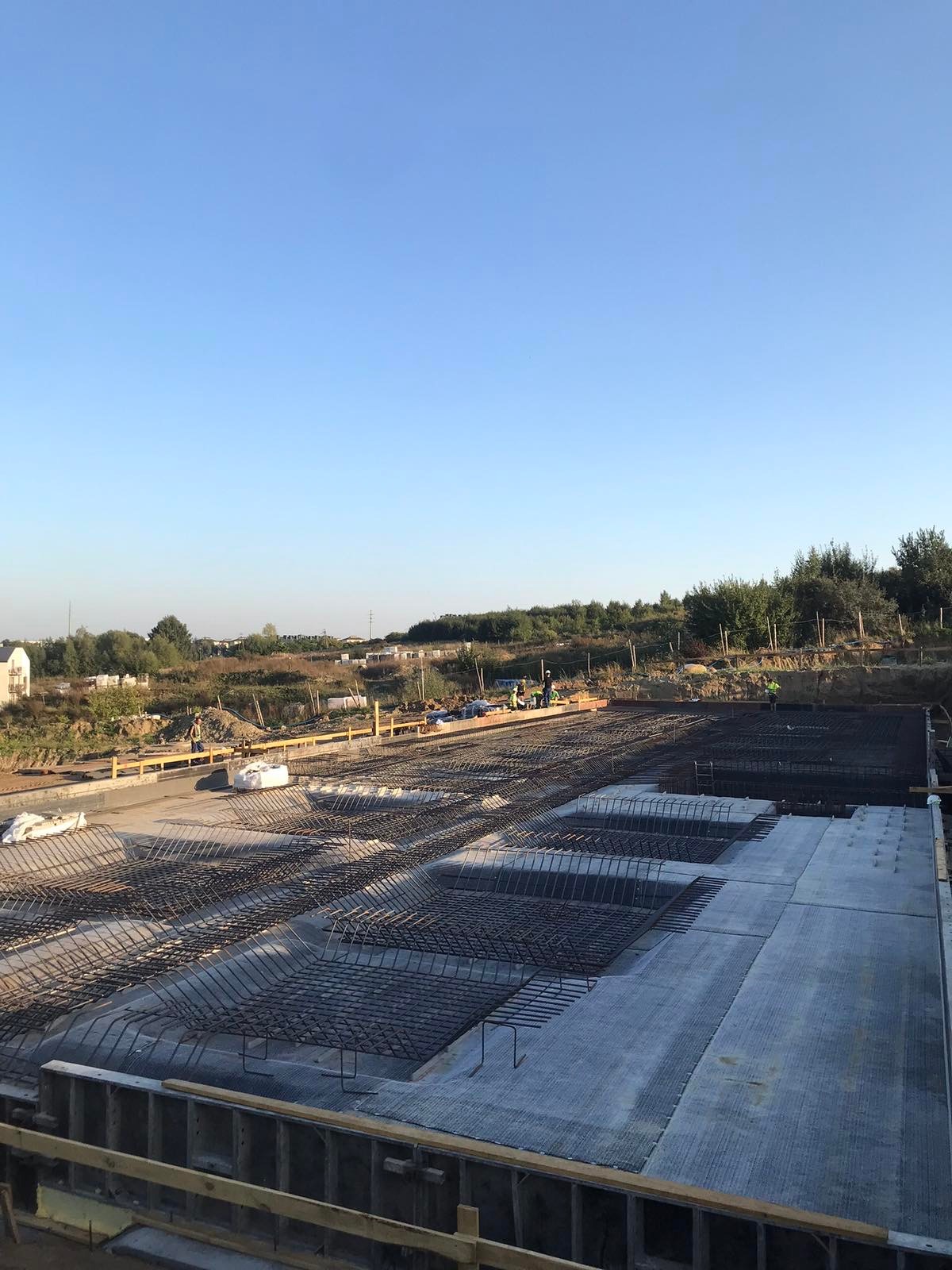 Przygotowanie zbrojenia i wylewki betonowej pod fundamenty budynku, widoczne szalunki i pracownicy w oddali.