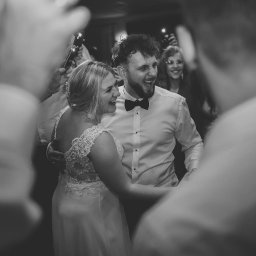 Julia Foto Olympia - Czarno-białe zdjęcie uśmiechniętej pary młodej w tańcu, otoczonej przez gości weselnych z telefonami. Panna młoda w koronkowej sukni, pan młody w muszce.