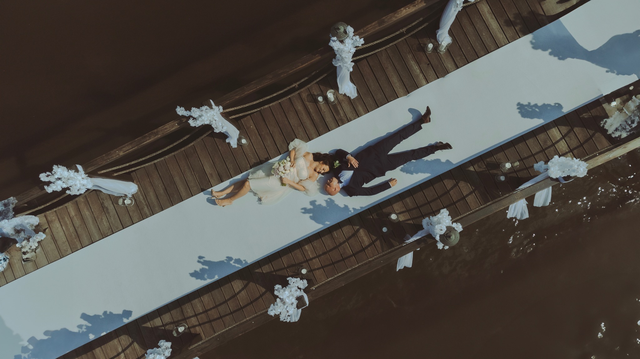 Para młoda leżąca na białym dywanie na drewnianym pomoście udekorowanym białymi kwiatami i świecami, widok z góry, romantyczna sesja.