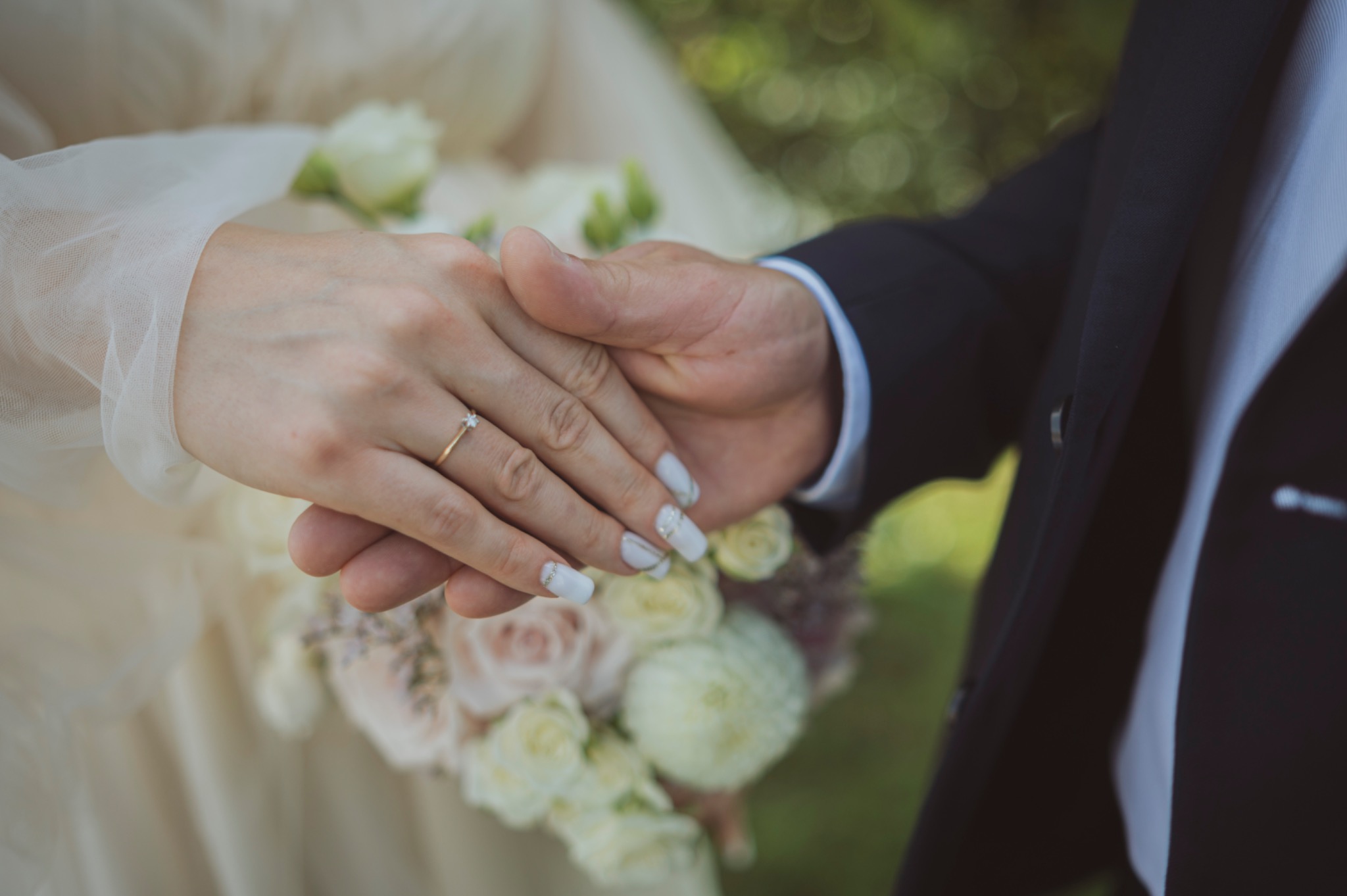 Dłonie pary młodej z obrączką na tle bukietu ślubnego, Chrzanów. Ona w białej sukni, on w garniturze. Delikatny manicure panny młodej.