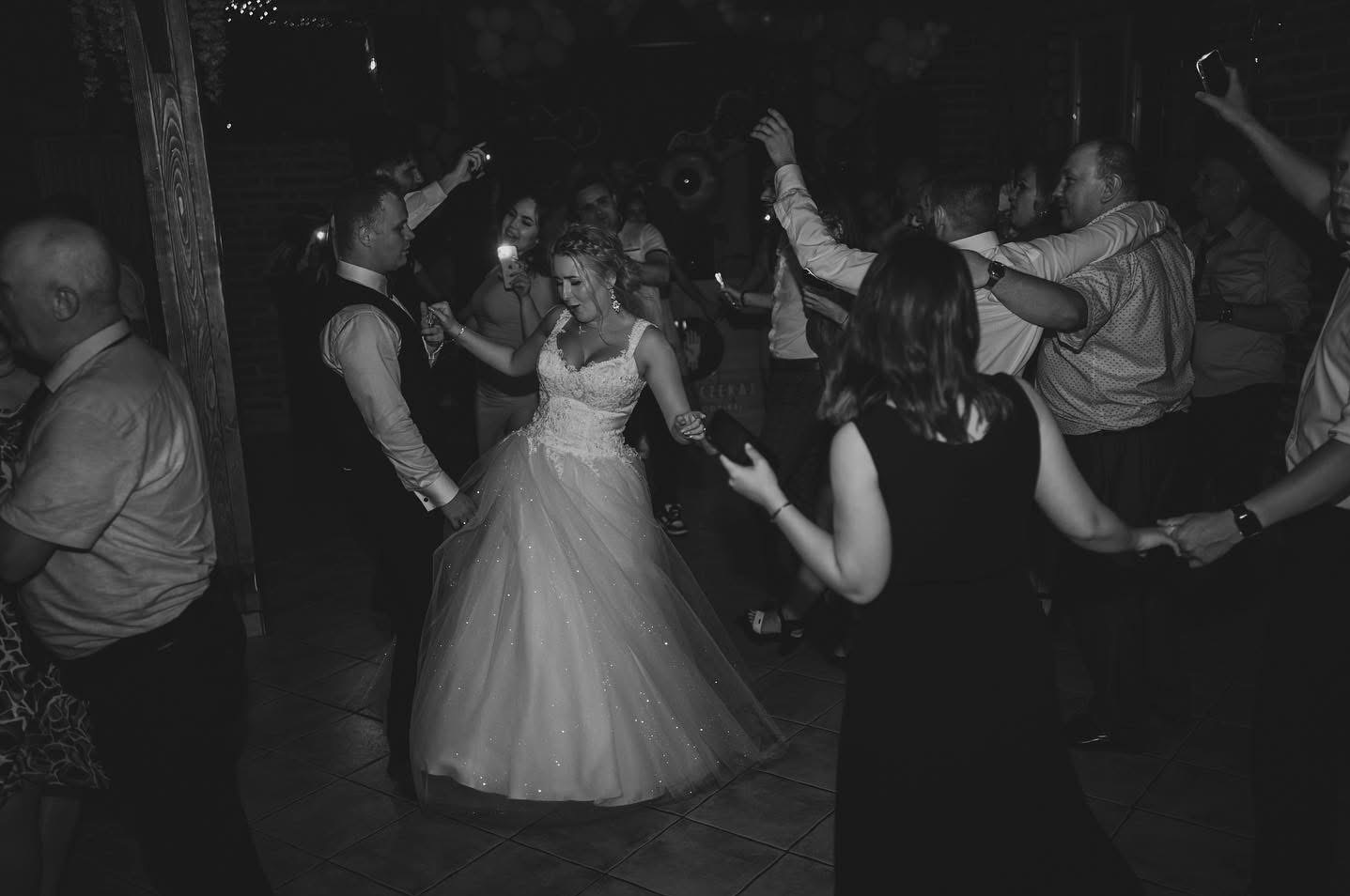 Czarno-białe zdjęcie tańczącej pary młodej w otoczeniu gości z telefonami. Ujęcie z lekkiej perspektywy, światło podkreśla suknię panny młodej.