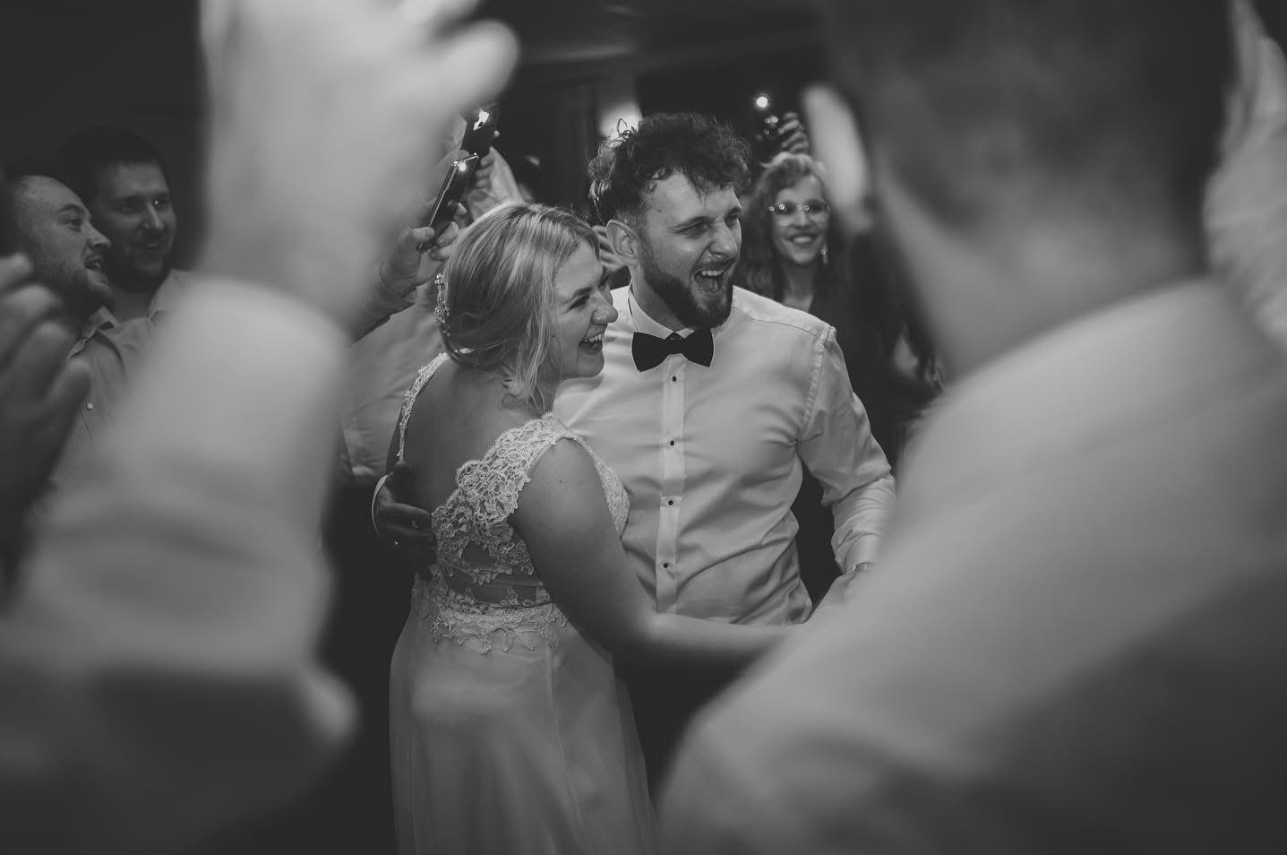 Czarno-białe zdjęcie uśmiechniętej pary młodej w tańcu, otoczonej przez gości weselnych z telefonami. Panna młoda w koronkowej sukni, pan młody w muszce.