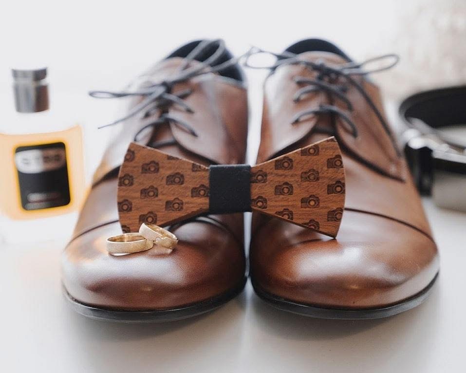 Brązowe, skórzane buty z drewnianą muszką w aparaty i obrączkami. W tle flakon perfum. Detal ślubny w ciepłej tonacji, oświetlenie naturalne.