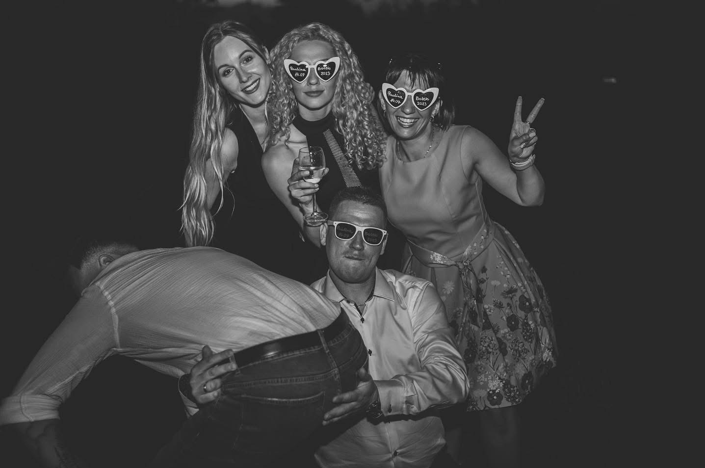 Czarno-białe zdjęcie grupy osób na imprezie, dwie kobiety w okularach w kształcie serc z napisem, mężczyzna w okularach i koszuli, śmiejący się i pozujący do zdjęcia.