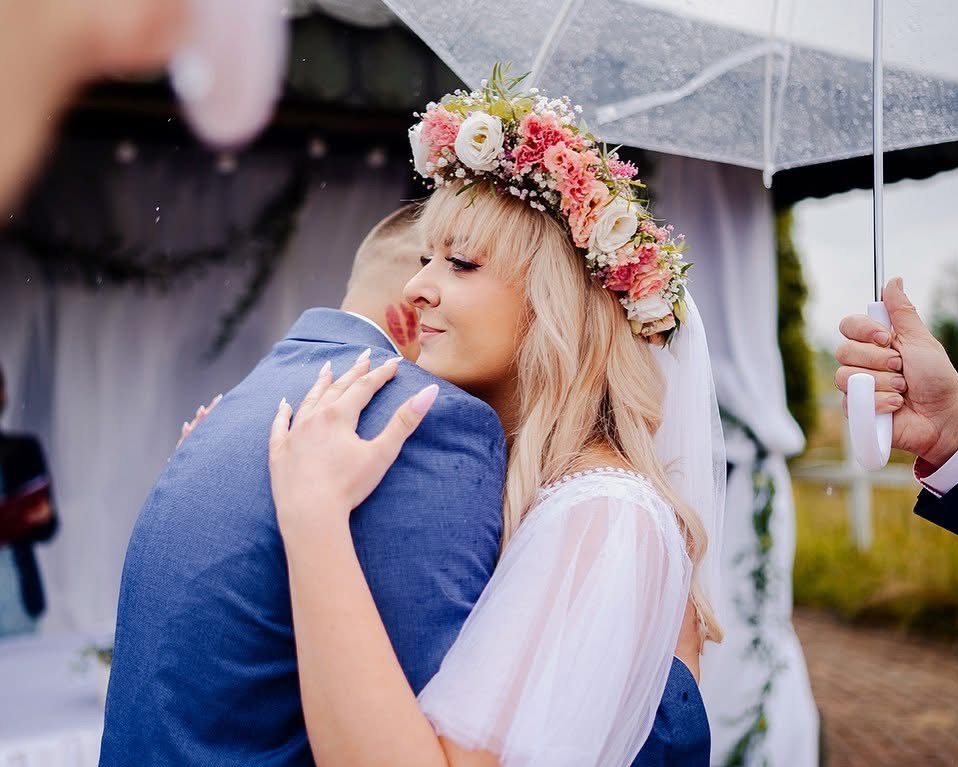 Panna młoda w wianku z kwiatów i welonie tuli pana młodego w niebieskim garniturze. Na policzku pana młodego widoczny ślad szminki. W tle dekoracje i parasol.