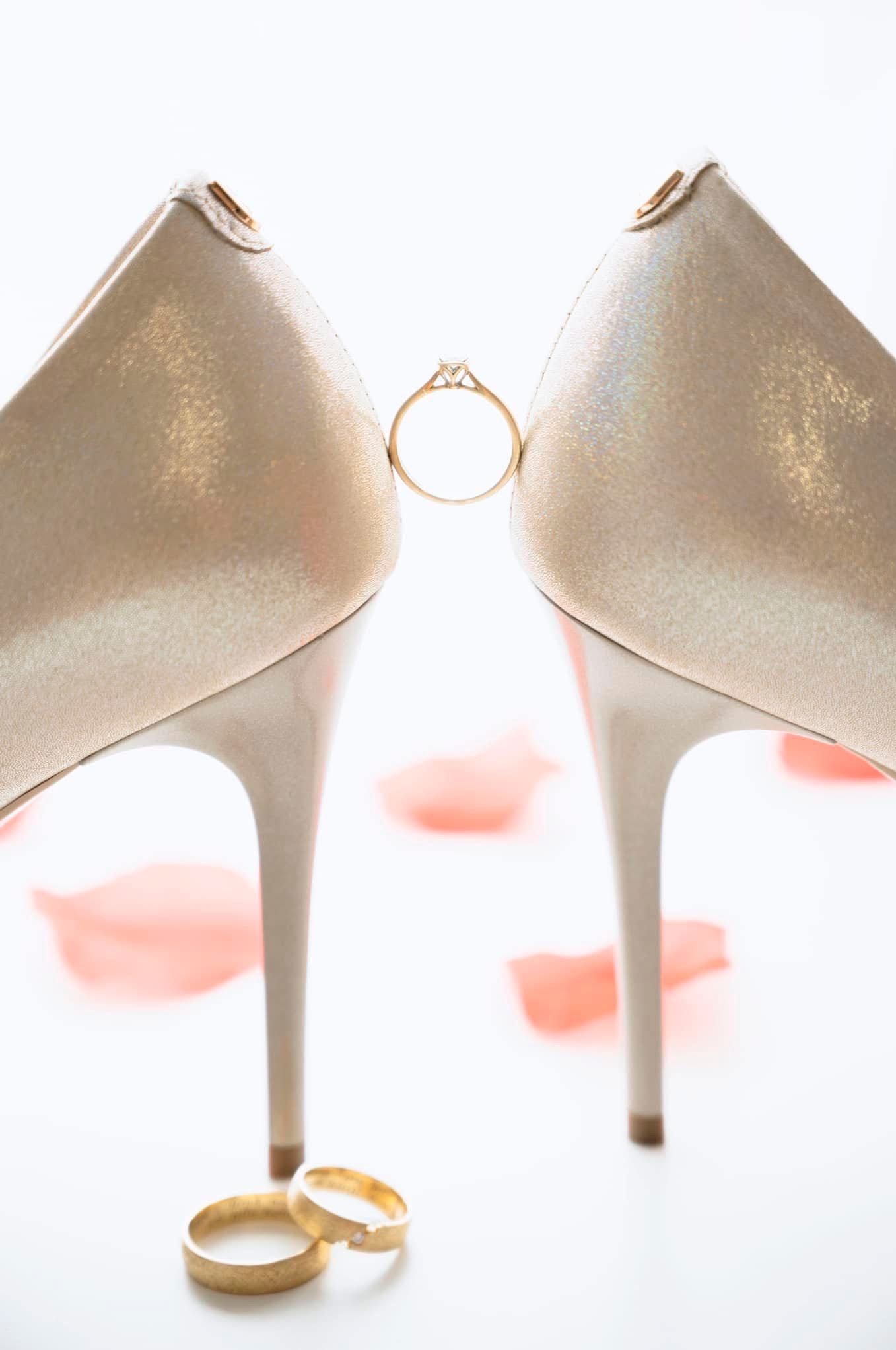 Dwa złote pierścionki leżące obok szpilek z umieszczonym pomiędzy nimi pierścionkiem zaręczynowym na tle rozrzuconych płatków róż.