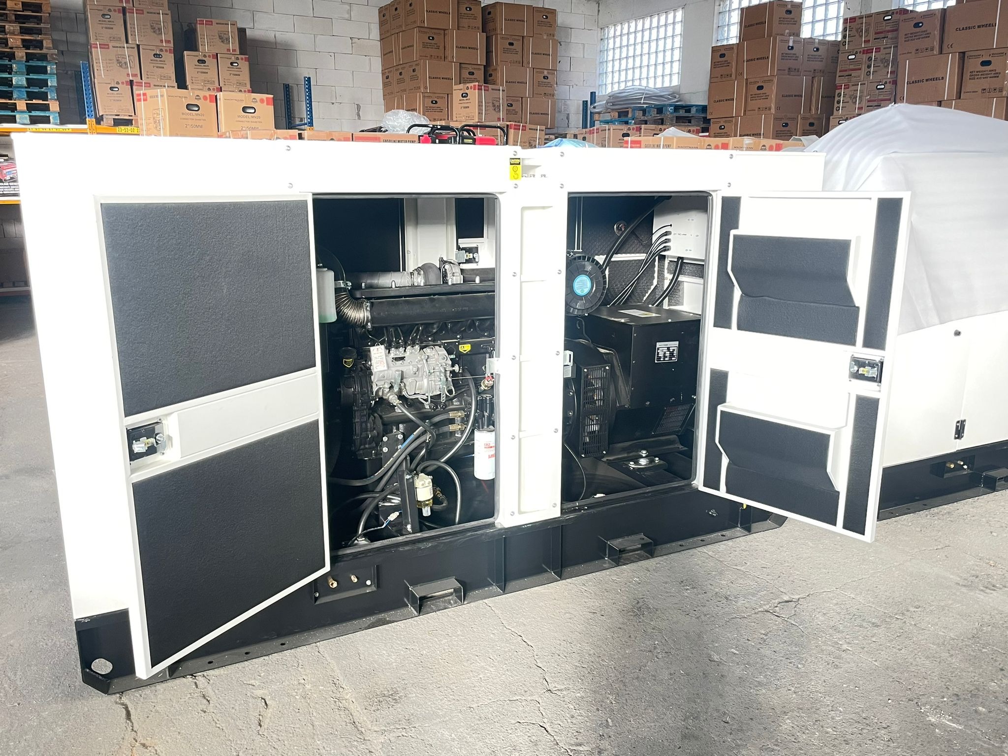 Otwarty agregat prądotwórczy w magazynie, widoczne silnik spalinowy i podzespoły elektryczne, kartony w tle.