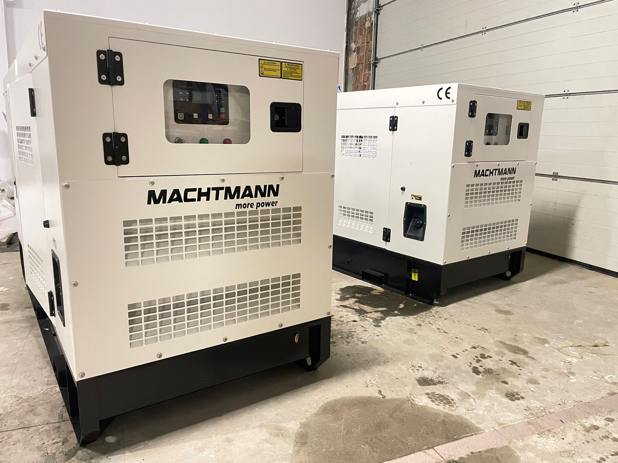 Dwa białe generatory marki Machtmann z widocznymi panelami sterowania, ustawione w magazynie z betonową podłogą i bramą rolowaną w tle.