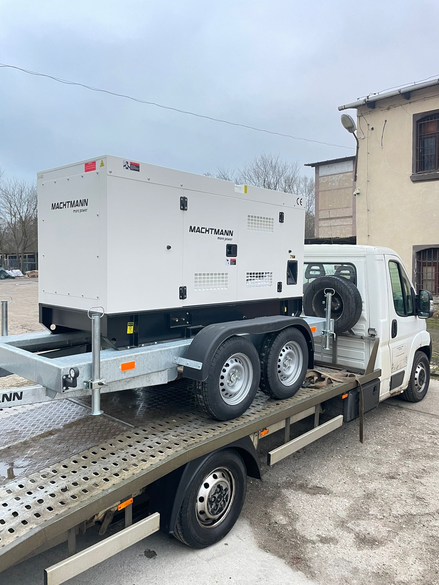 Agregat prądotwórczy marki Machtmann umieszczony na platformie transportowej za białym samochodem dostawczym, na tle budynku z widoczną lampą uliczną.