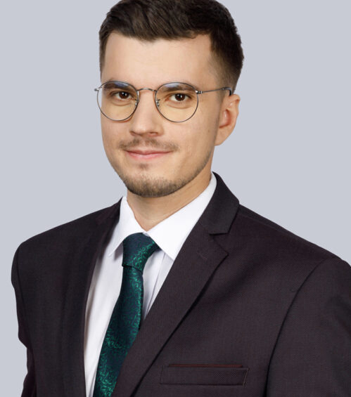 Portret mężczyzny w okularach i ciemnym garniturze z zielonym krawatem na jednolitym, jasnoszarym tle.