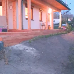 Nowo wybudowany drewniany taras przy domu jednorodzinnym, widoczny dmuchawa do liści na betonowych schodach i częściowo przygotowany teren pod ogród.