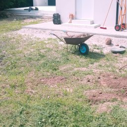 GardenComplex - Przygotowanie terenu pod ogród: pracownik z łopatą kopie ziemię, obok taczka z ziemią, grabie i paleta z kostką brukową w tle.