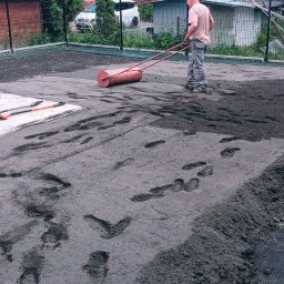 GardenComplex - Mężczyzna wyrównuje teren wałem ogrodowym, widoczne ślady stóp na świeżo rozplantowanej ziemi, obok ściany budynku leżą dwie szpadle, w tle siatka ogrodzeniowa i zabudowania.