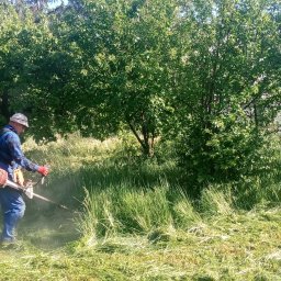 GardenComplex - Mężczyzna w kombinezonie i kapeluszu, z założonymi czerwonymi rękawicami, przycina wysoką trawę za pomocą podkaszarki spalinowej, w tle widoczne drzewa.