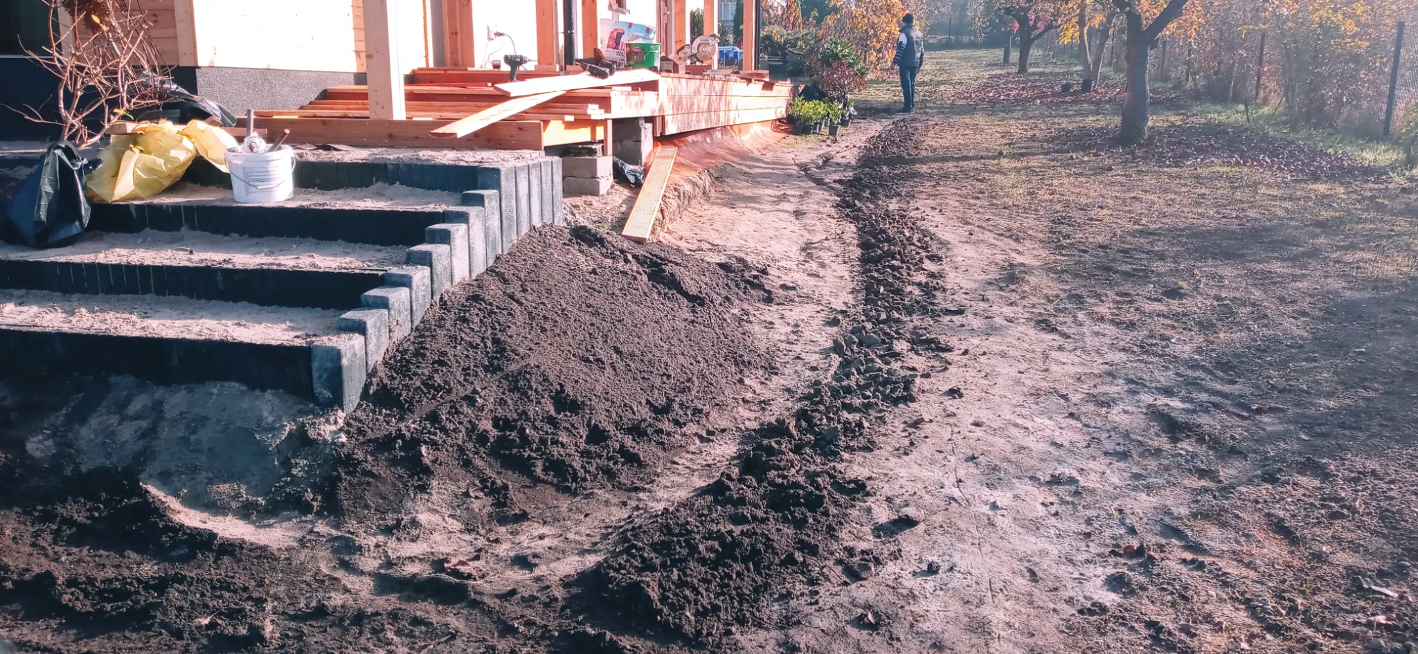 Przygotowanie terenu pod budowę tarasu i schodów z betonowych elementów, widoczny wykopany rów i pryzma ziemi, w tle osoba na trawniku i drzewa.