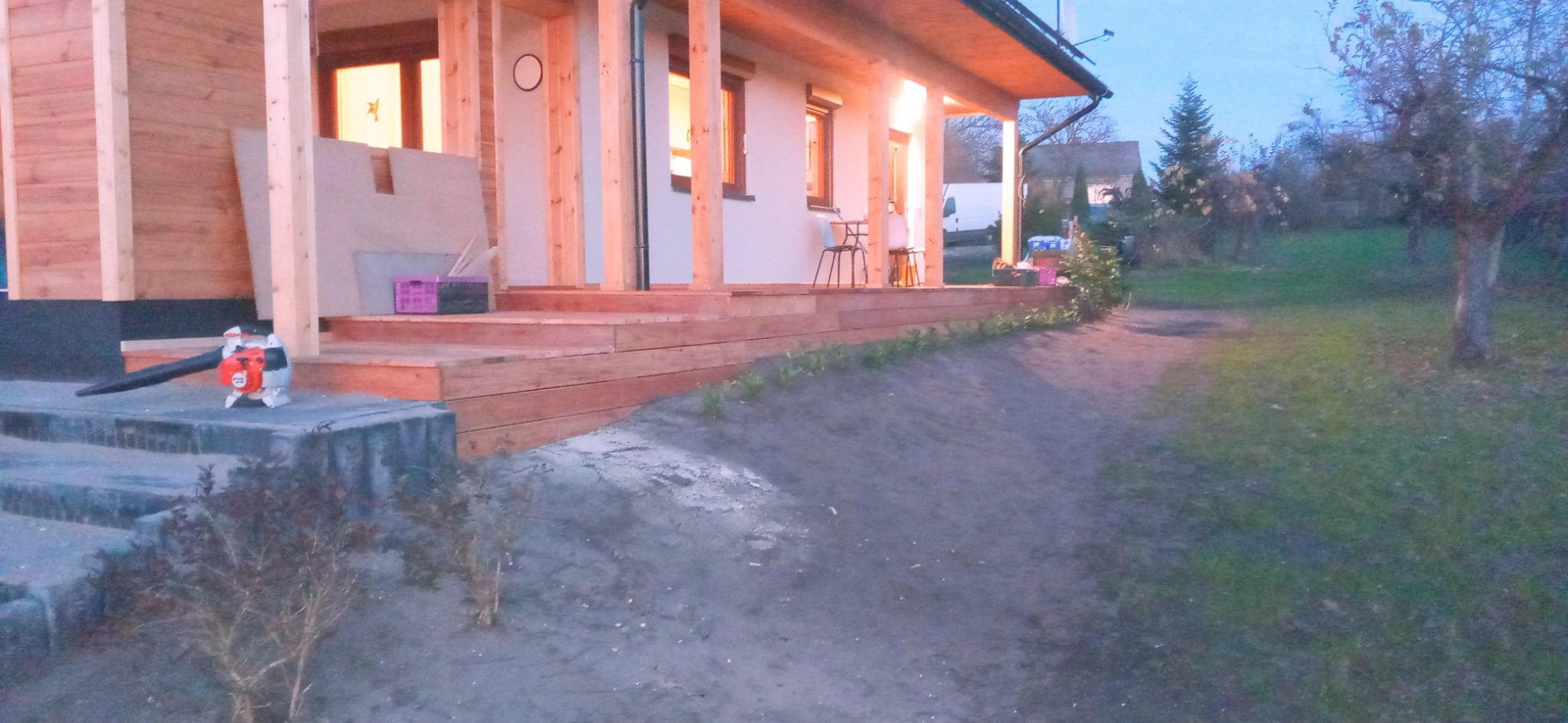 Nowo wybudowany drewniany taras przy domu jednorodzinnym, widoczny dmuchawa do liści na betonowych schodach i częściowo przygotowany teren pod ogród.