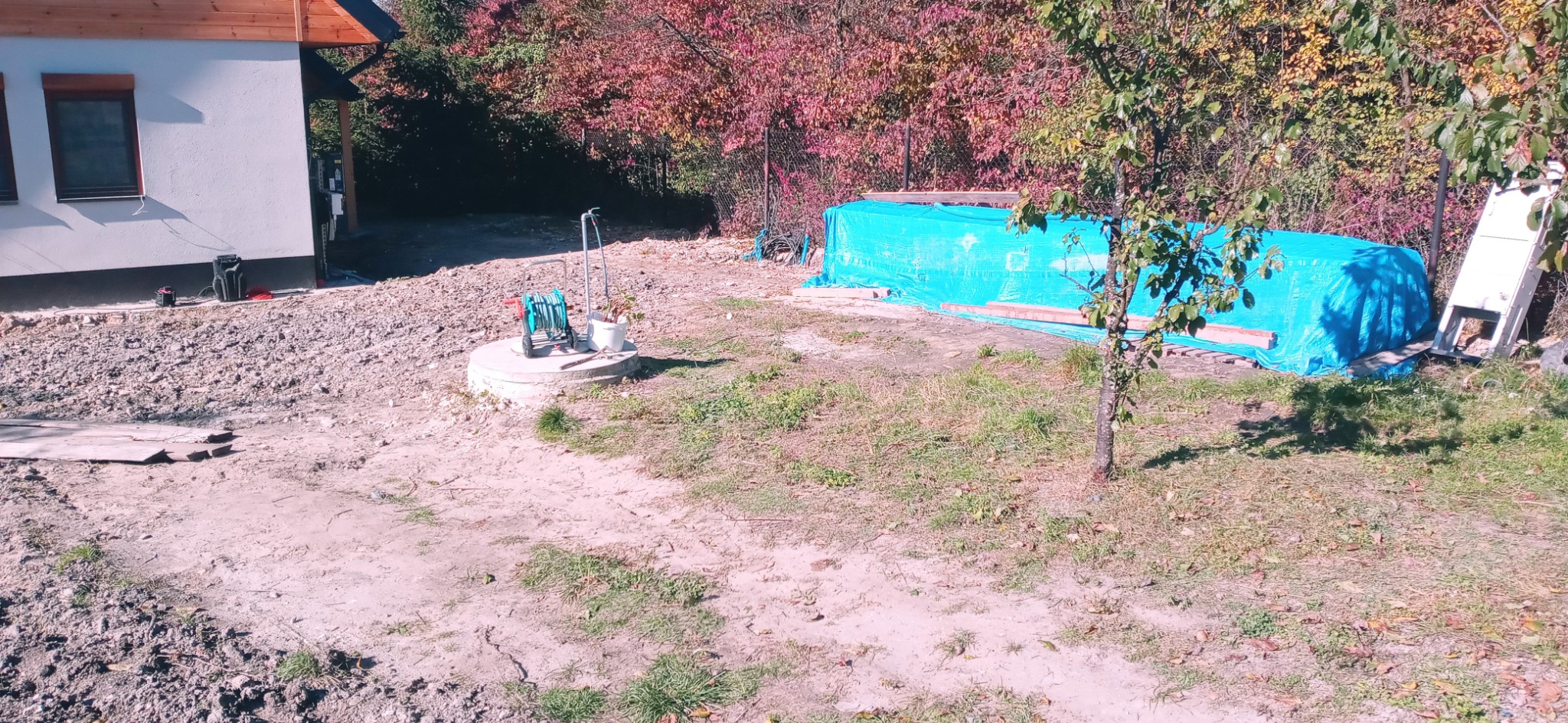 Przygotowany teren pod przyszły ogród z widocznym nowym domem, stertą ziemi przykrytą plandeką, studzienką z wężem ogrodowym i młodym drzewkiem.