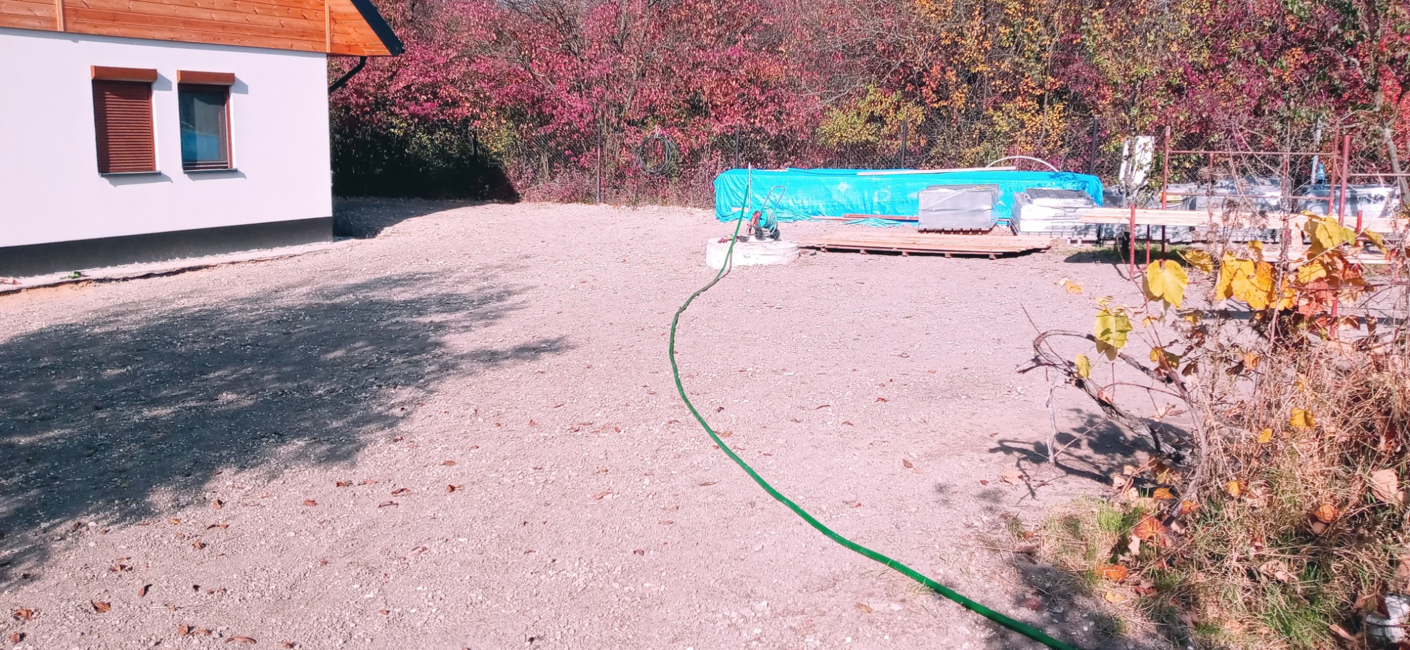 Teren przygotowany pod przyszły ogród z widocznymi materiałami budowlanymi pod plandeką, zielonym wężem ogrodowym i fragmentem budynku z brązowymi okiennicami.