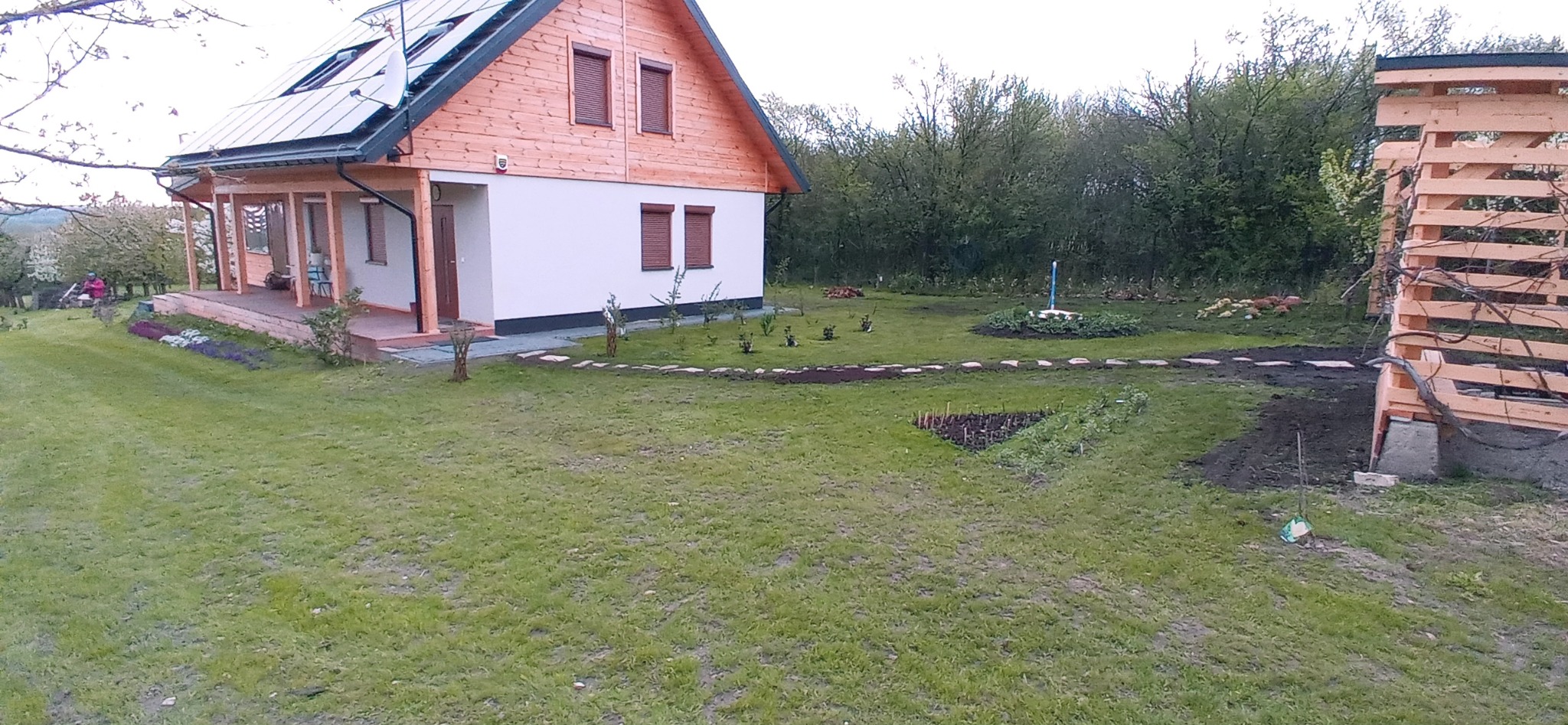 Rozległy ogród z nowo posadzonymi drzewkami, krzewami i rabatami, częściowo wyłożony kamieniami, z widocznym drewnianym domem jednorodzinnym w tle oraz konstrukcją z drewnianych belek po prawej...