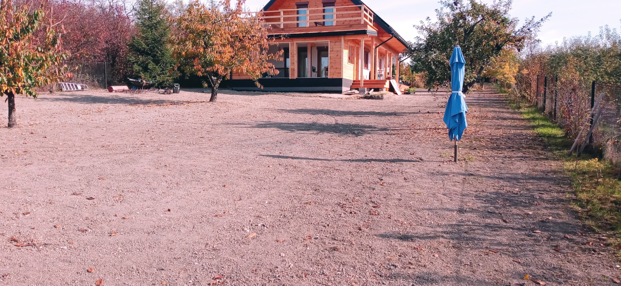 Widok na drewniany dom z ogrodem w trakcie zakładania, z widoczną niezrównaną ziemią i złożonym niebieskim parasolem.