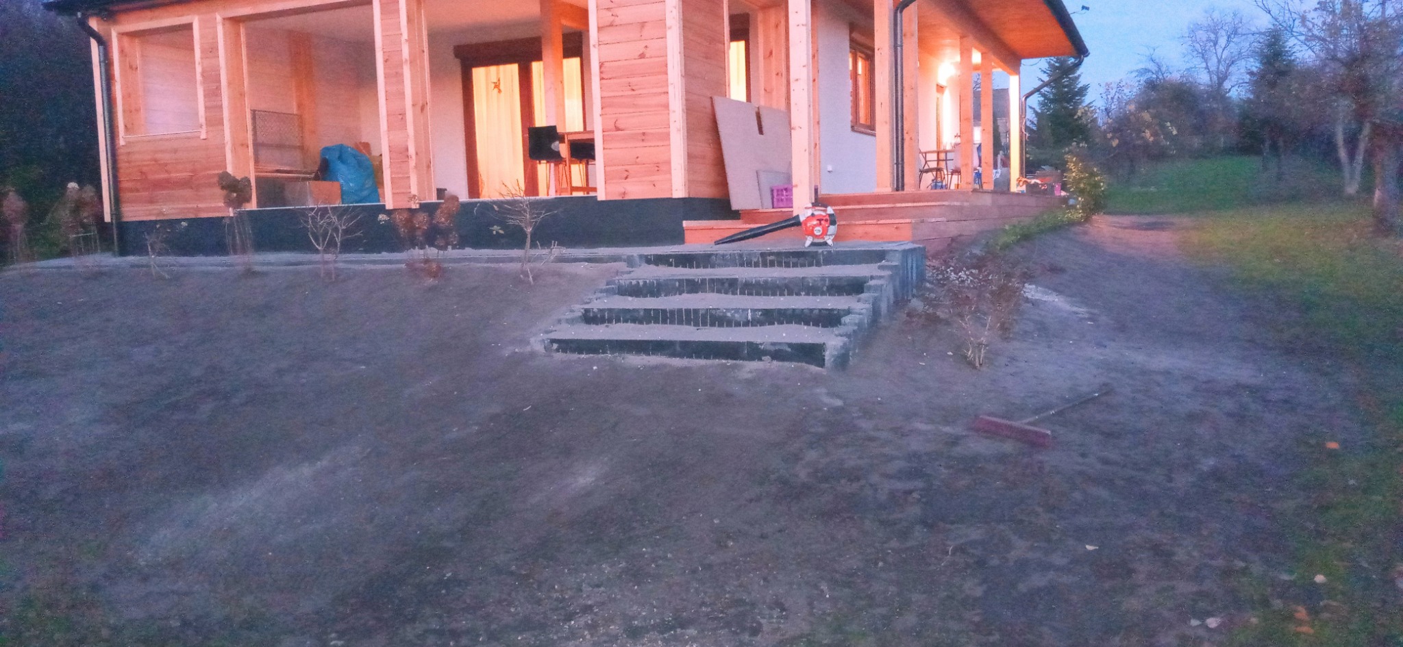 Nierówny teren z nowo posadzonymi krzewami, betonowe schody prowadzące do drewnianego domu, dmuchawa do liści na schodach, grabie leżące na ziemi.