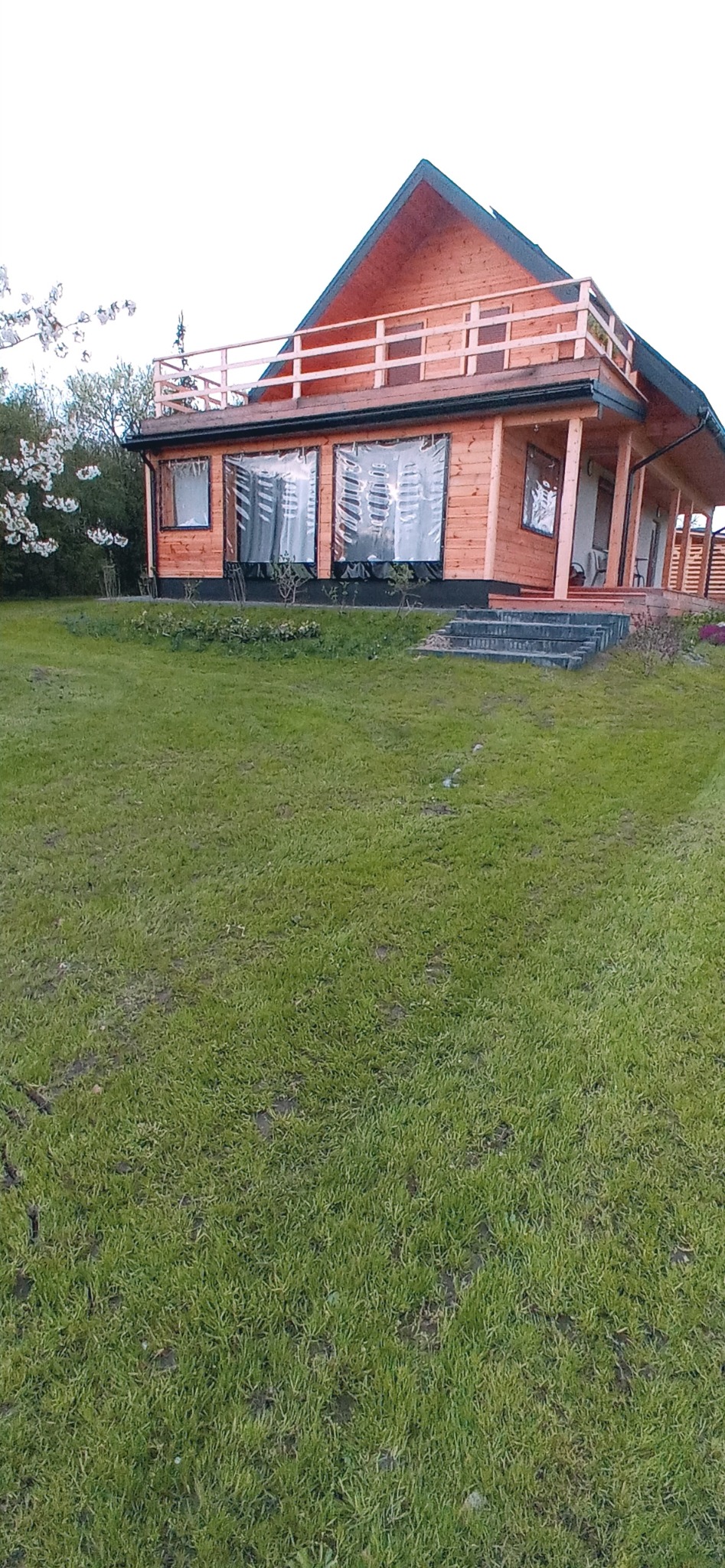 Drewniany dom z balkonem na dachu, otoczony trawnikiem i młodymi drzewkami.